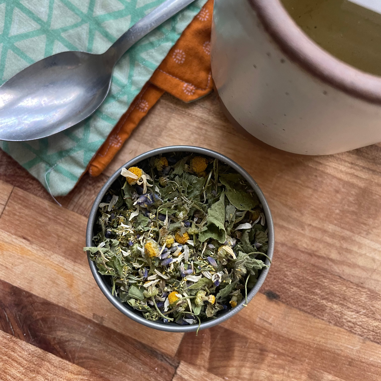 A small metal bowl filled with loose herbal tea sits on a wooden table, alongside a spoon on a green patterned cloth and a ceramic mug containing brewed tea.