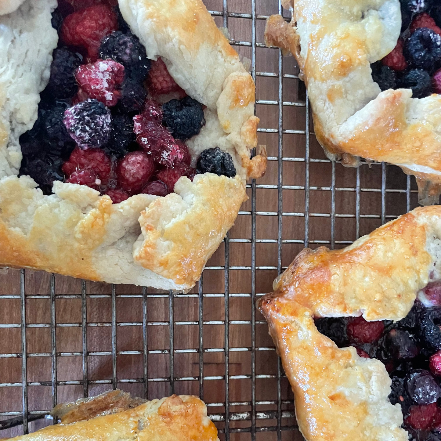 Close-up of several baked berry galettes with golden flaky crusts and mixed berries, arranged on a metal cooling rack over a wooden surface.
