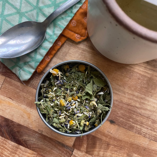 A small metal bowl filled with loose herbal tea sits on a wooden table, alongside a spoon on a green patterned cloth and a ceramic mug containing brewed tea.