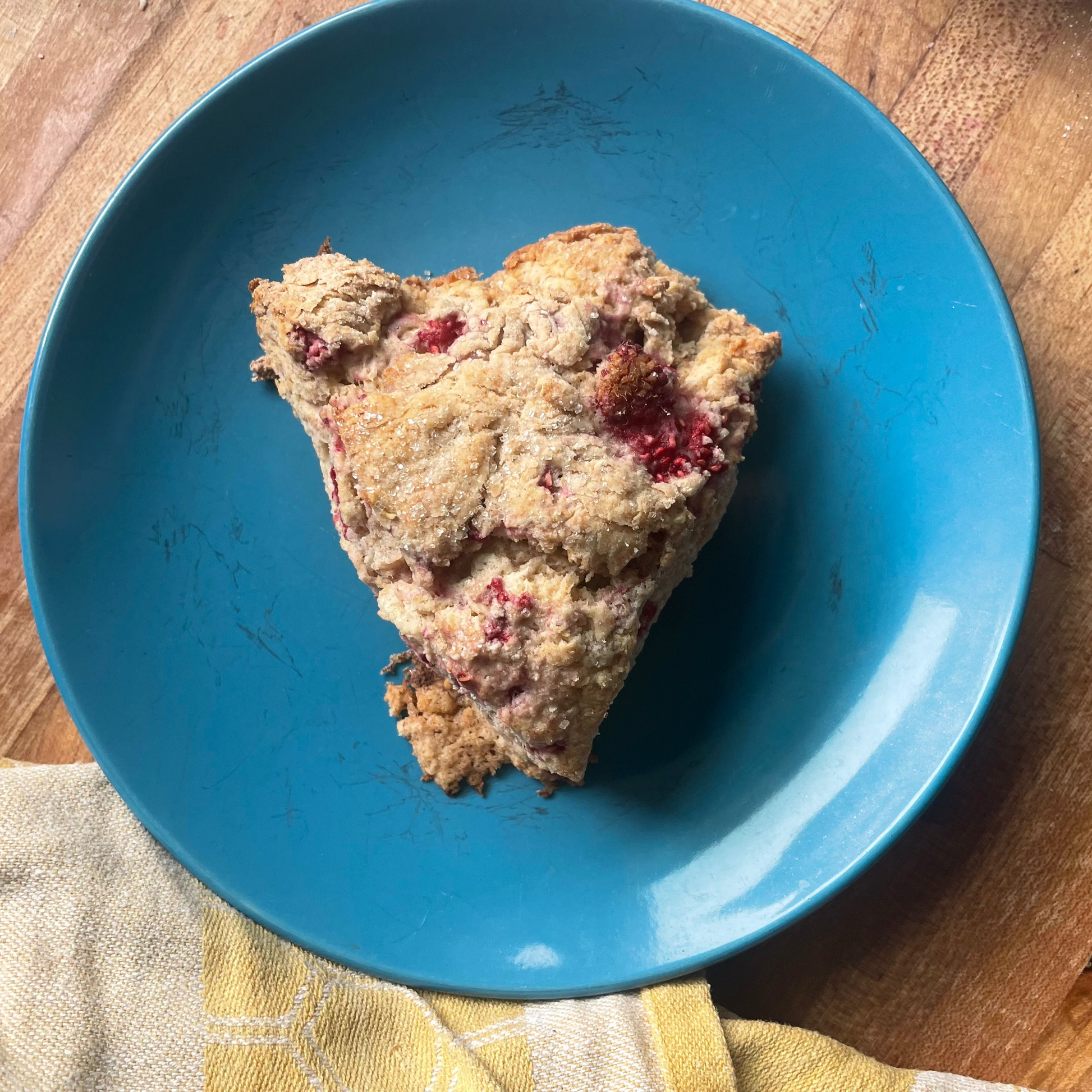 A triangular slice of baked scone or biscuit with visible red fruit pieces, served on a blue plate over a wooden surface with a yellow cloth.