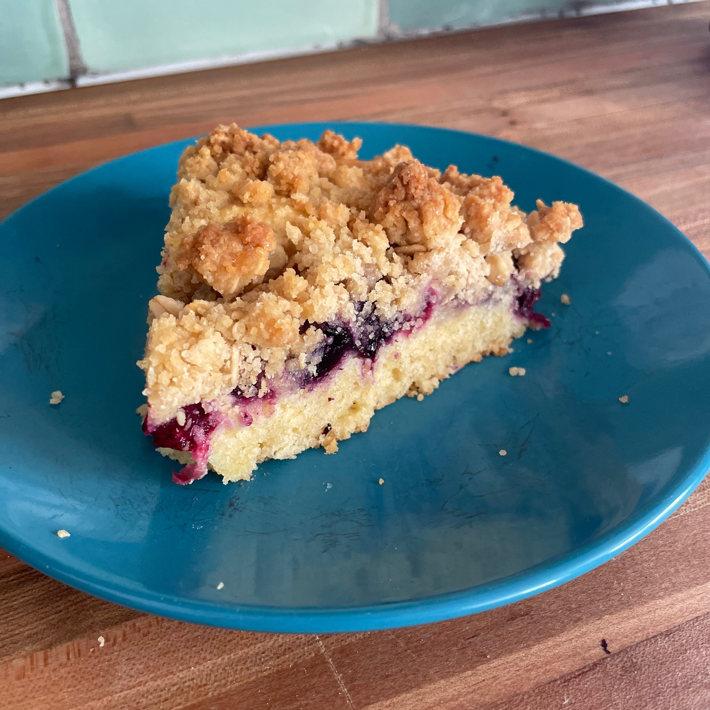 A slice of blueberry crumb pie served on a blue plate on a wooden table with a tiled wall in the background.